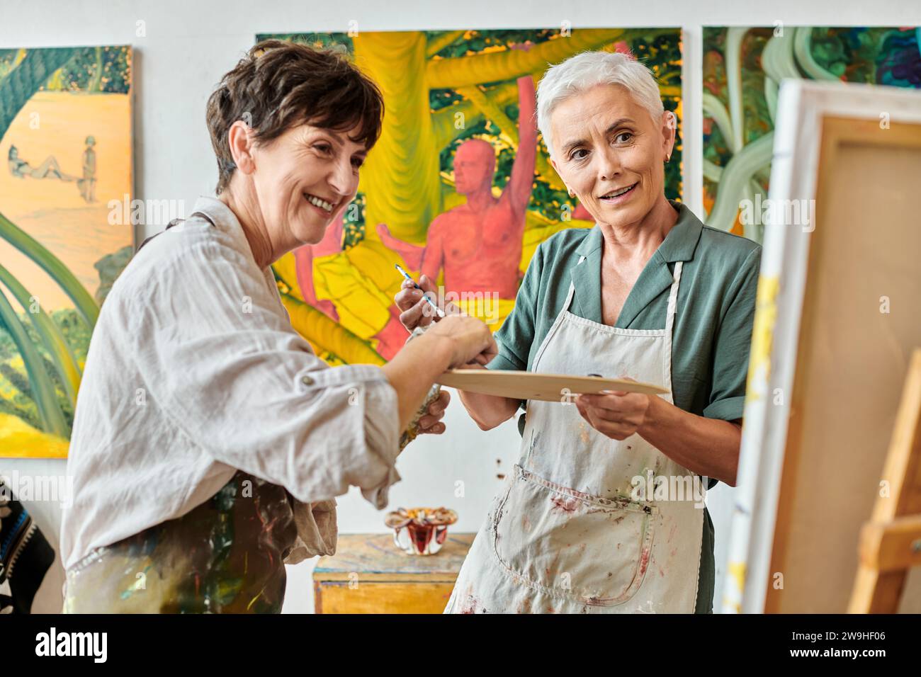 Lächelnde reife Frauen mit Farbpalette, die während des Meisterkurses im Kunststudio Staffelei betrachten Stockfoto