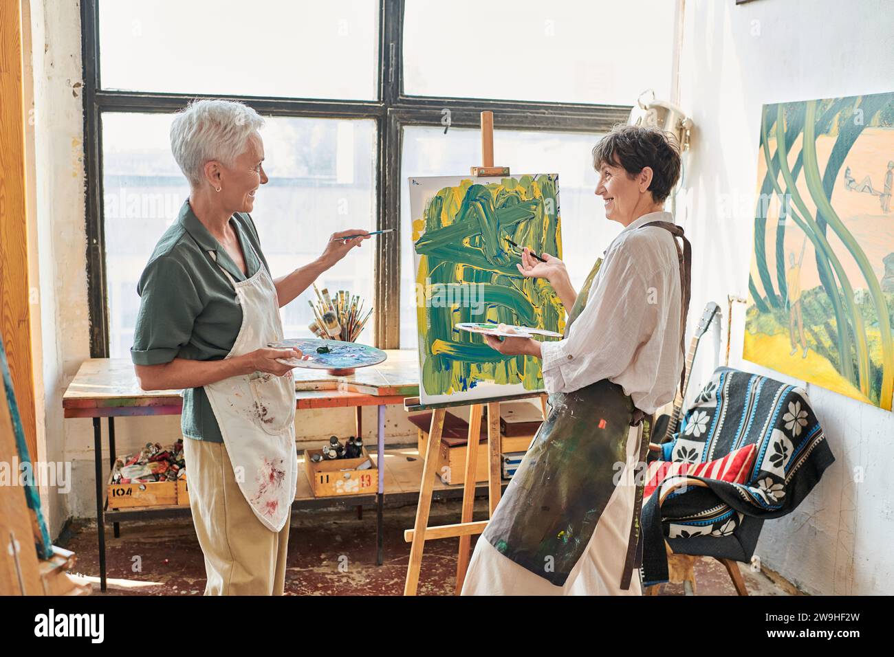 Fröhliche Frau, die auf Staffelei zeigt mit bunten Gemälden in der Nähe einer Freundin im Kunststudio, Teamwork Stockfoto