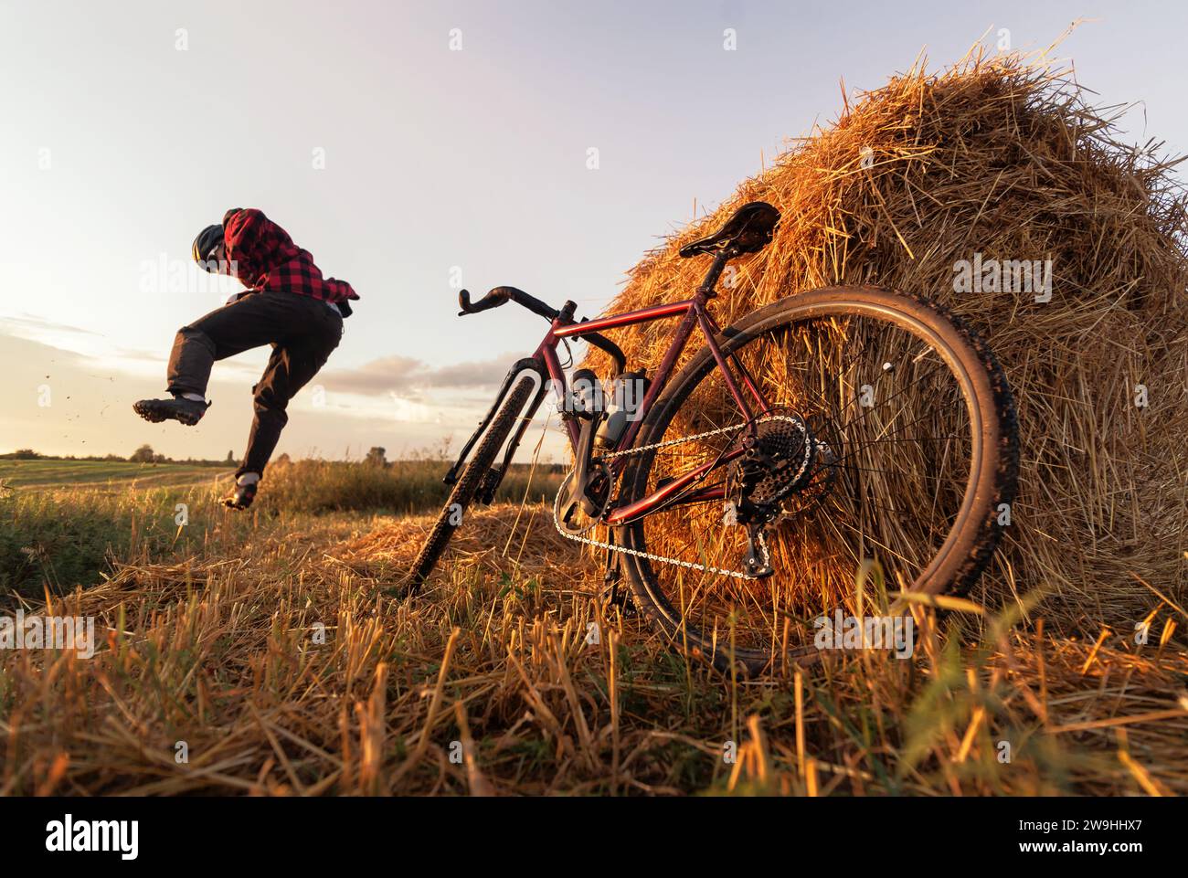 Glücklicher Radfahrer, der in einem Feld mit Heuhaufen und einem Fahrrad in der Nähe bei Sonnenuntergang springt. Aktives Radtraining. Stockfoto
