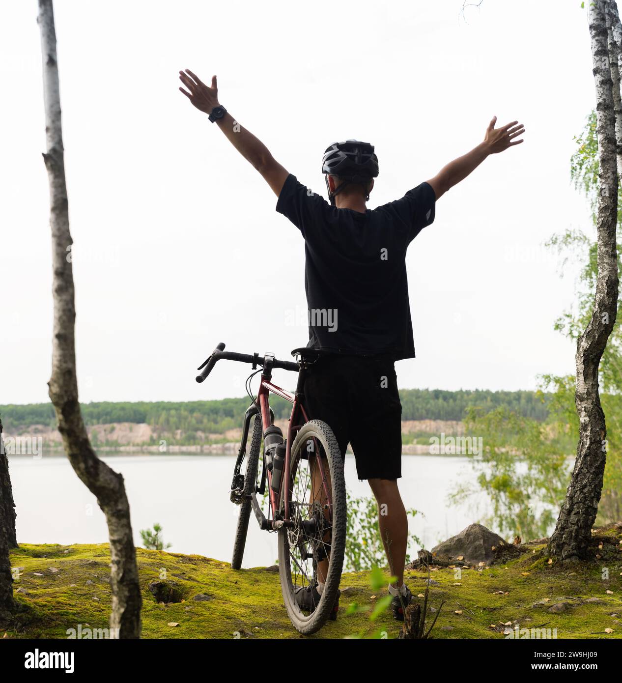 Radfahrer mit erhobenen Armen in einem Helm steht an einer Klippe über dem See. Konzept von Erfolg, Sieg, Zielerreichung und Sieg. Glückliche Radfahrer machen Stockfoto