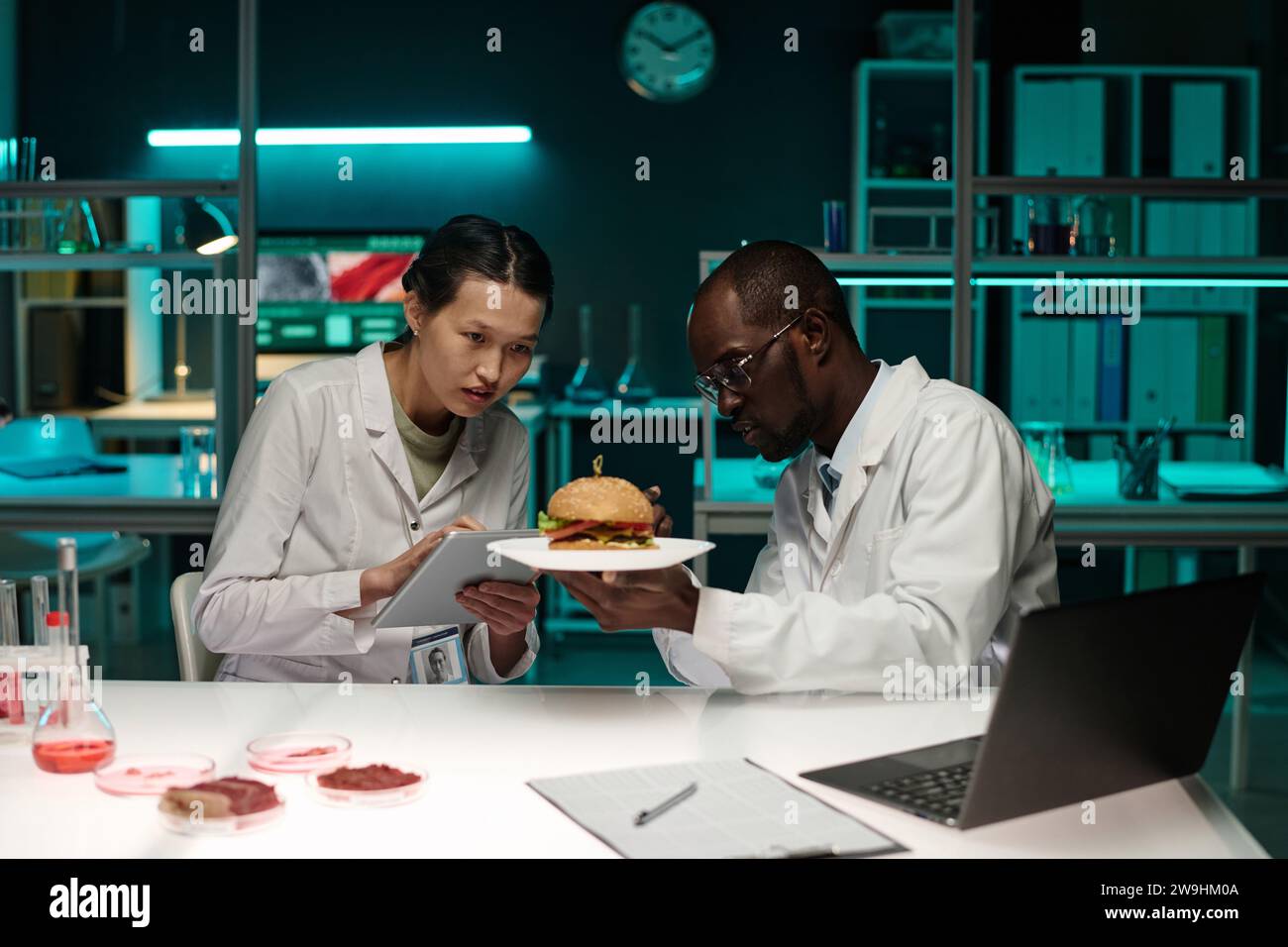 Zwei Wissenschaftler analysierten Hamburger mit Fleisch aus Labor, eine Forscherin macht Notizen auf Tablette Stockfoto