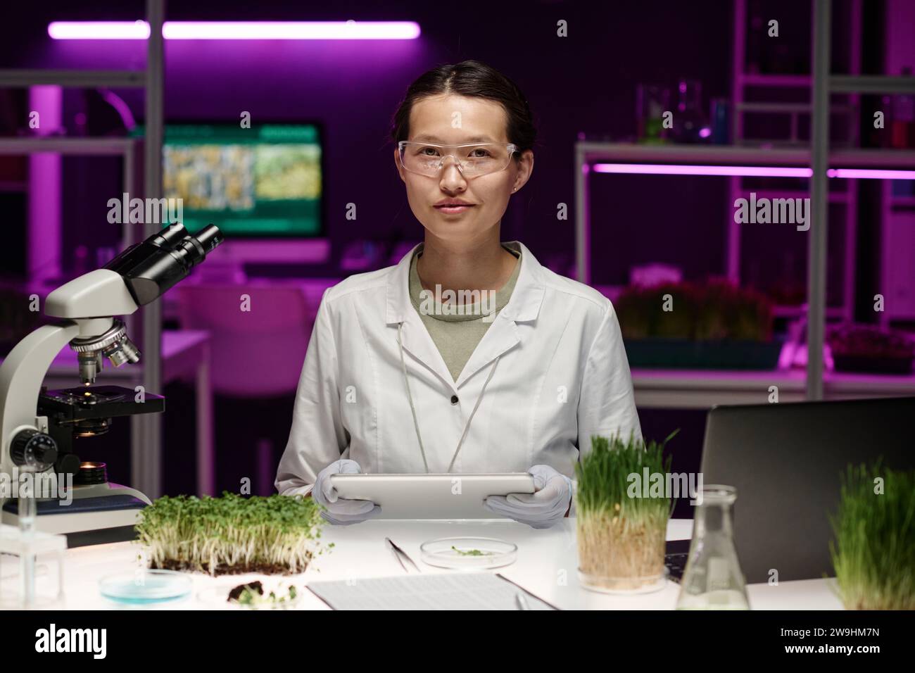 Mittlere Aufnahme einer asiatischen Laborarbeiterin, die mit Ausrüstung an ihrem Schreibtisch sitzt und in die Kamera schaut Stockfoto