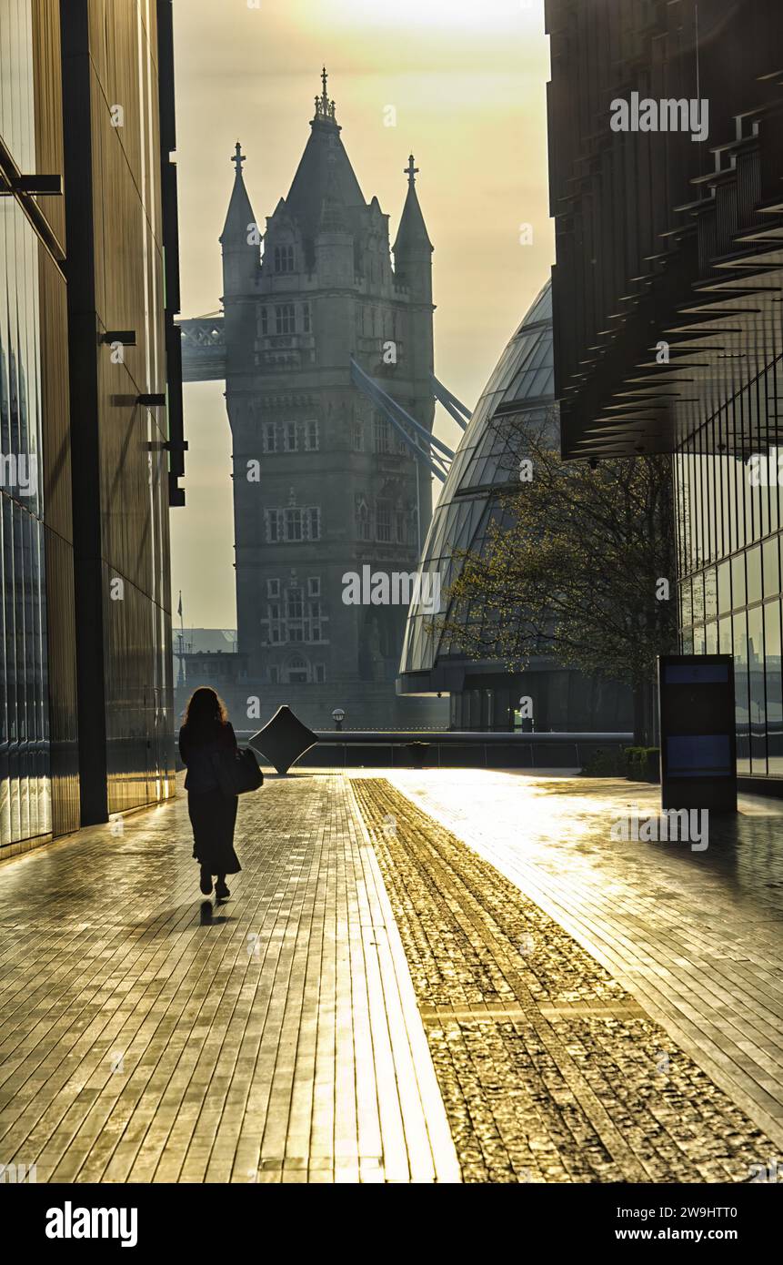 Silhouette einer Frau, die auf dem Bürgersteig zwischen Gebäuden mit einem Turm der Tower Bridge im Hintergrund in die Morgensonne geht Stockfoto