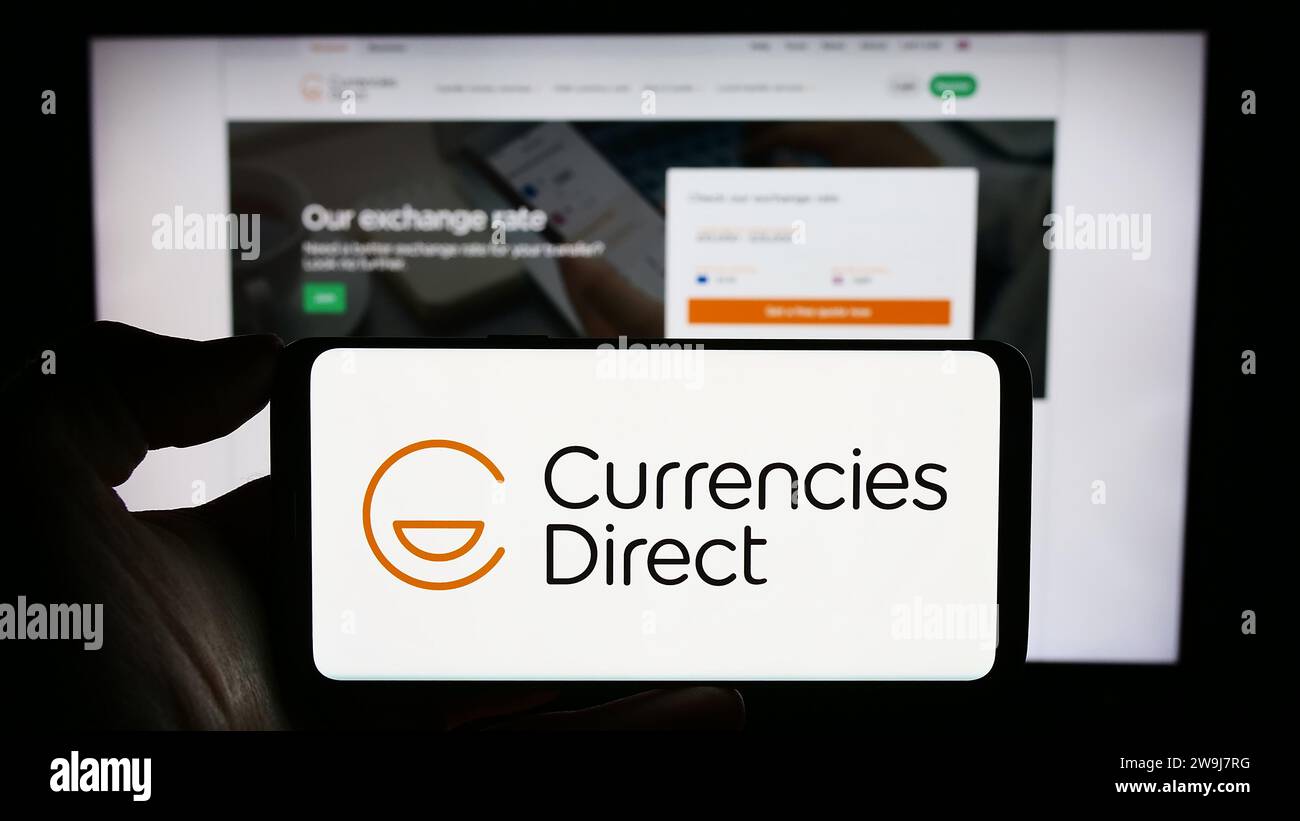 Person, die ein Mobiltelefon mit dem Logo der britischen Geldtransfergesellschaft Currency Direct Ltd. Vor der Webseite des Unternehmens hält. Schwerpunkt auf dem Display des Telefons. Stockfoto
