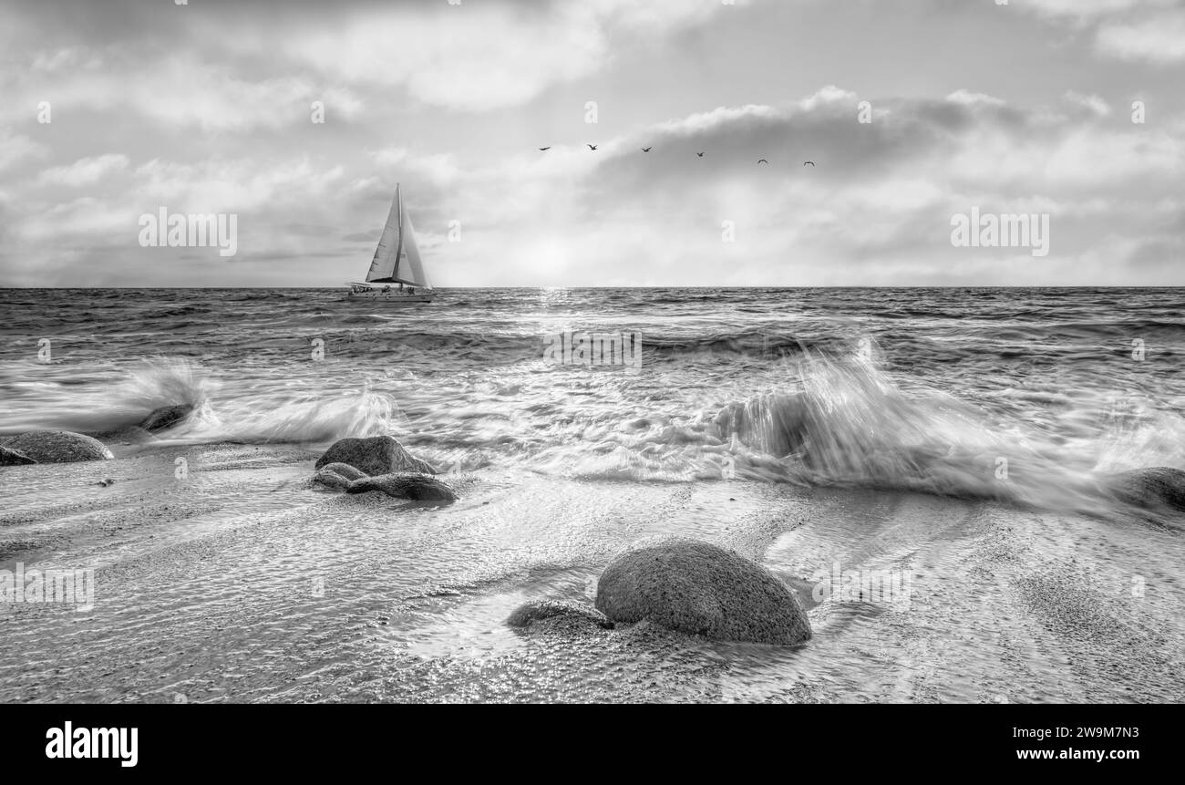 Ein Segelboot segelt entlang des Ozeans mit Wellenbrechern auf Shore Black and White Stockfoto