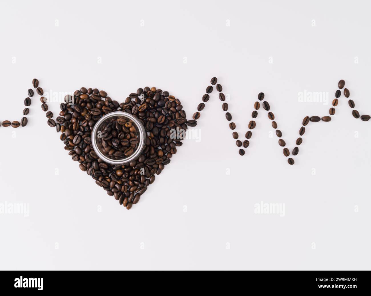 Kreatives Layout mit Herzform aus gerösteten Kaffeebohnen und Kaffeetasse, das die Beziehung zwischen Kaffeetrinken und Herz-Kreislauf-Erkrankungen zeigt. Stockfoto