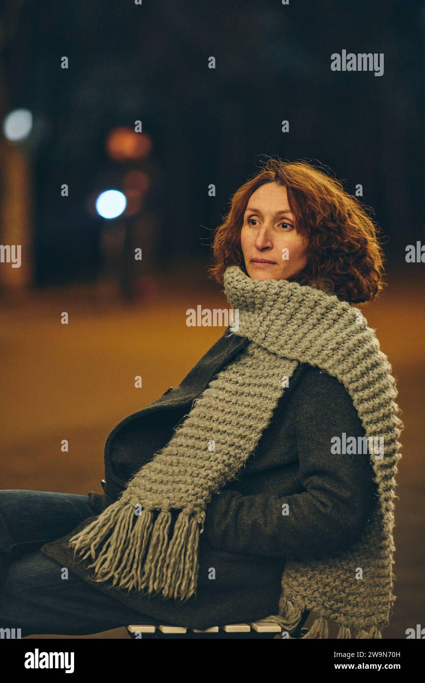Nachdenkliche Frau mittleren Alters, die in der Nachtstadt auf einem Stuhl sitzt Stockfoto