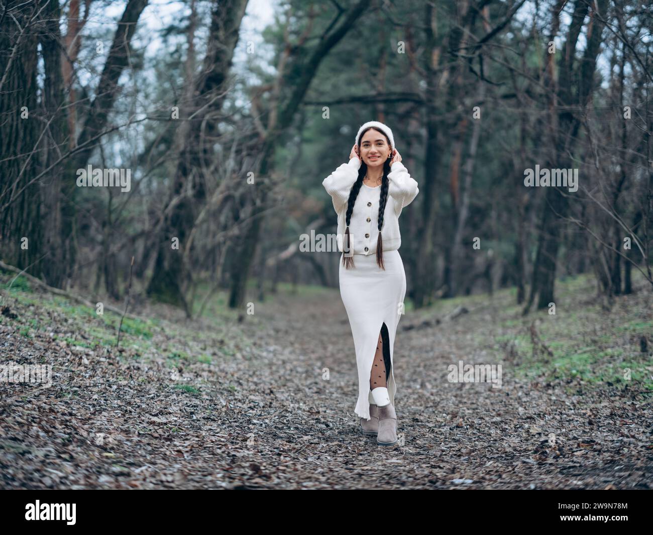 Glückliche, lächelnde junge Frau mit geflochtenen Haaren, die in weißem Pullover, Rock und Hut durch den Wald geht. Stockfoto