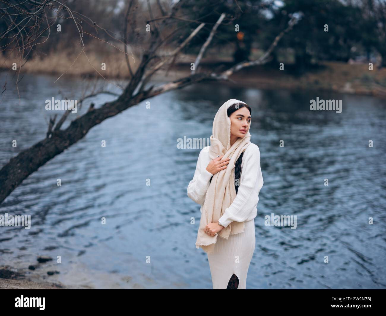 Junge Frau mit geflochtenen Haaren, die im Wald in weißem Pullover, Rock und Schal vor dem Hintergrund des Sees spaziert. Stockfoto