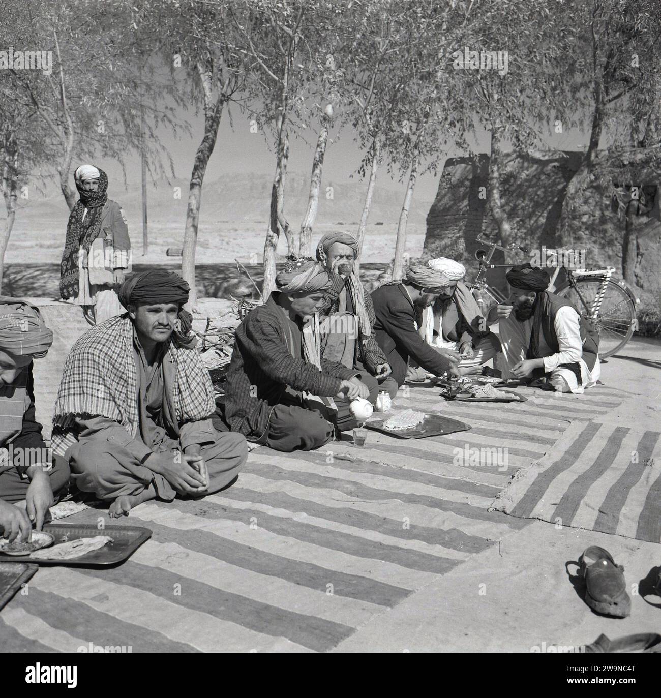 1950er Jahre, historische, männliche afghanen in traditioneller Kleidung, Kopfbedeckung und barfuß, sitzen auf Teppichen auf dem Boden draußen und essen, Afghanistan. Die Mehrheit der Afghanen lebt außerhalb der Städte in Stammesgemeinden, lebt traditionelle, bäuerliche llfestyles, in einer männlich dominierten, hoch patriarchalischen Kultur. Als größte ethnische Gruppe des Landes haben die Paschtunen strenge Ehrenregeln und die Auslegung des islamischen und der Scharia-Rechts. Stockfoto