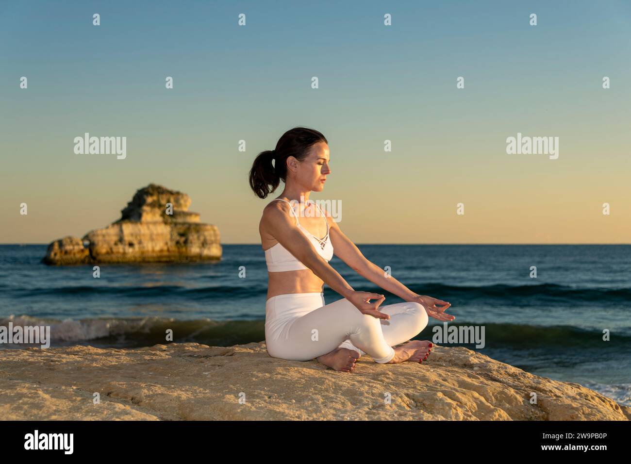 Frau, die auf einem Felsen am Meer sitzt und in der Sonne meditiert Stockfoto