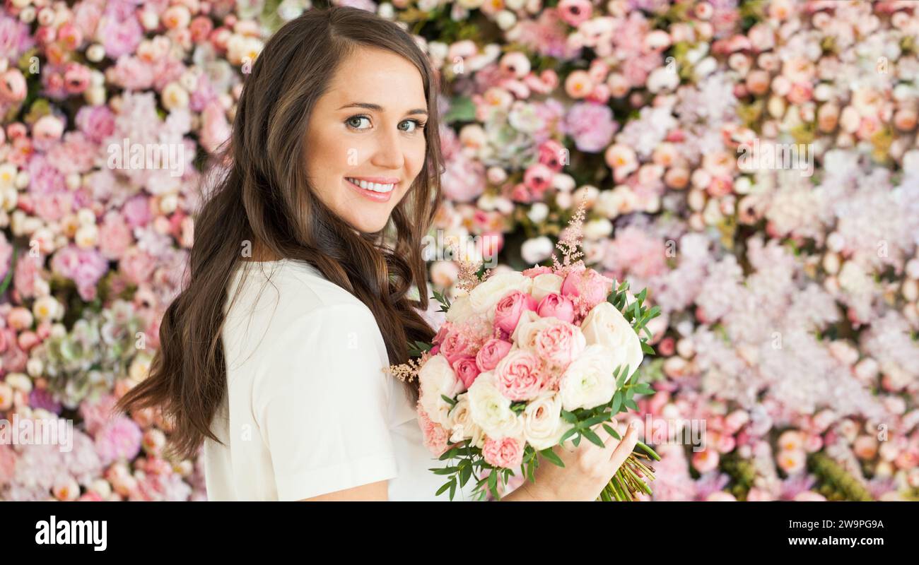 Hübsche glückliche brünette Frau mit niedlichem Lächeln, die Blumengeschenk hält und auf rosa floralem Hintergrund posiert. Blühend, Liebe, Geburtstagsfeierlichkeiten am 8. märz Stockfoto