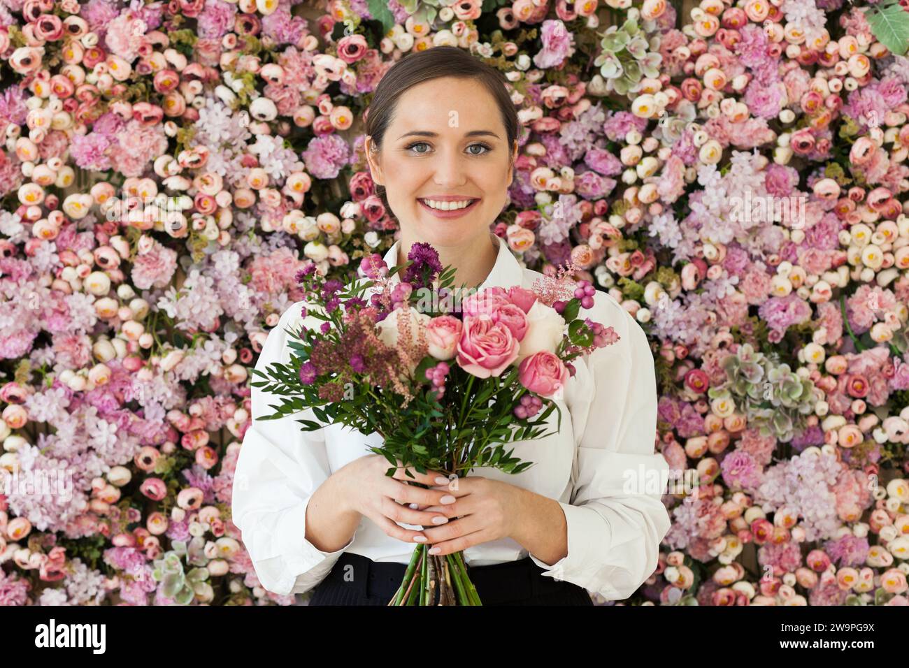 Glückliche lächelnde brünette Frau mit niedlichem Lächeln, die Blumengeschenk hält und auf rosa floralem Hintergrund posiert. Blühend, Liebe, Geburtstagsfeiertag am 8. märz Stockfoto