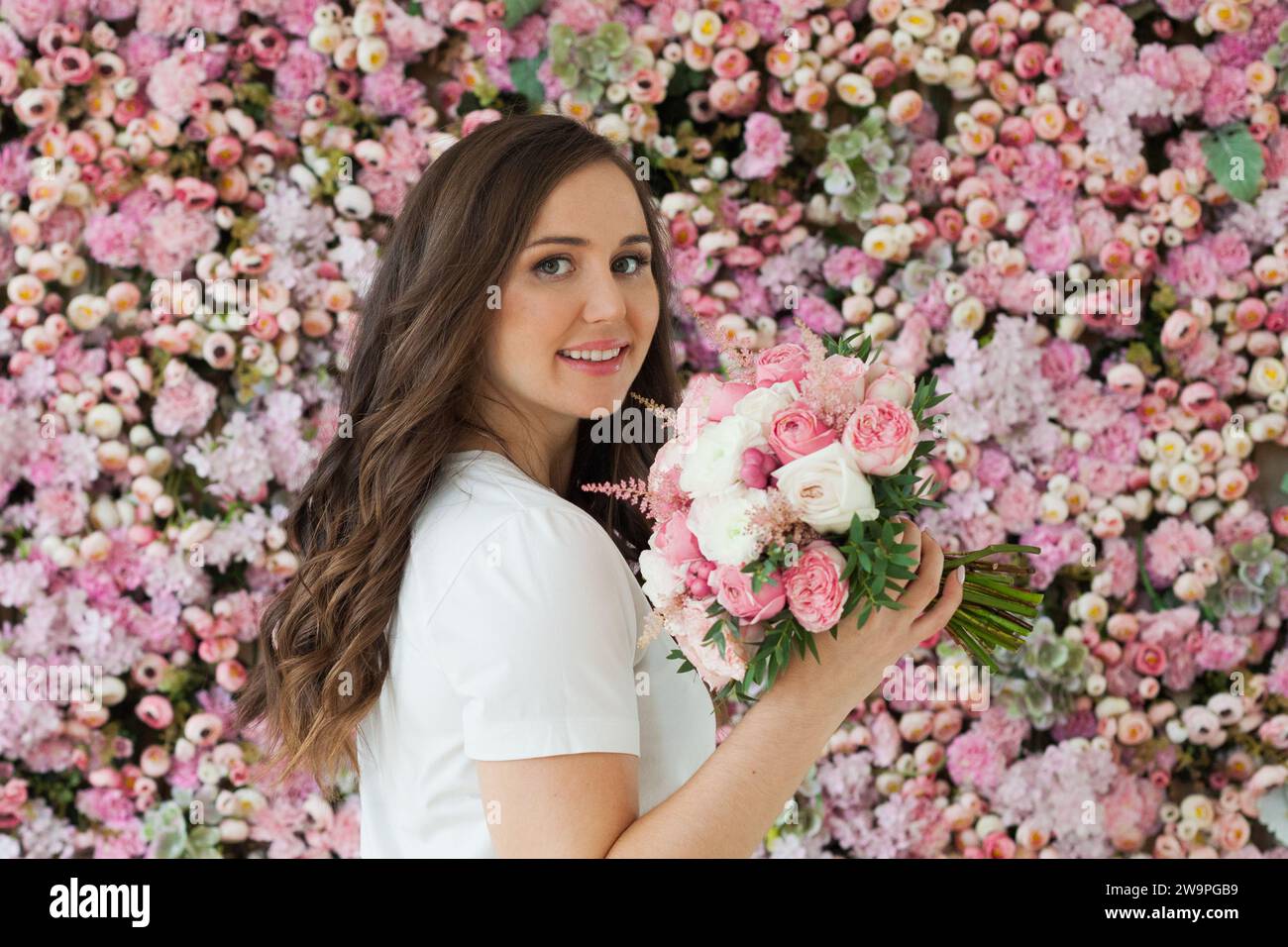 Nette glückliche Brünette Frau mit niedlichem Lächeln, die Blumengeschenk hält und auf rosa floralem Hintergrund posiert. Blooming, Liebe, Geburtstagskonzept am 8. märz Stockfoto