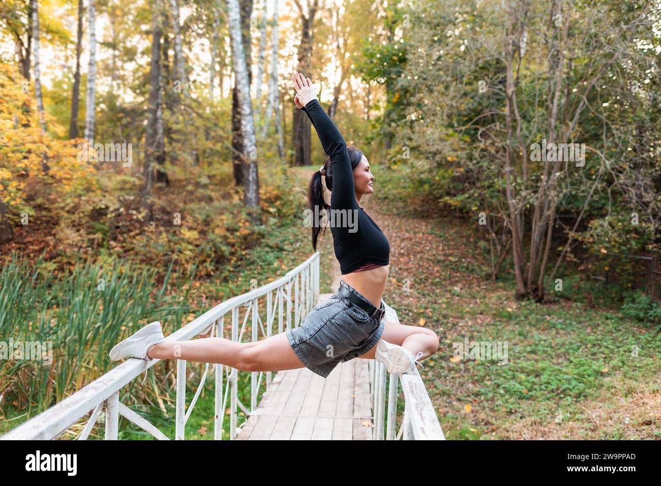 Attraktive Frau, die eine Variation der Eka Pada Rajakapotasana-Übung durchführt, Taubenpose, sitzt auf dem Geländer einer hölzernen Fußgängerbrücke Stockfoto