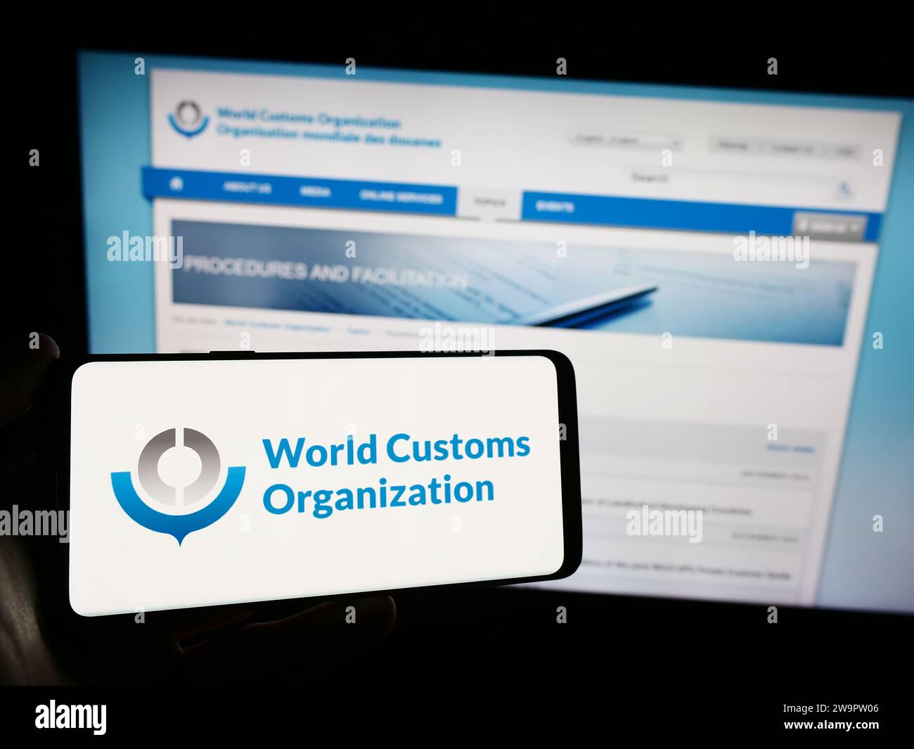 Person, die ein Mobiltelefon mit dem Logo der Weltzollorganisation (WZO) vor der Webseite hält. Schwerpunkt auf dem Display des Telefons. Stockfoto