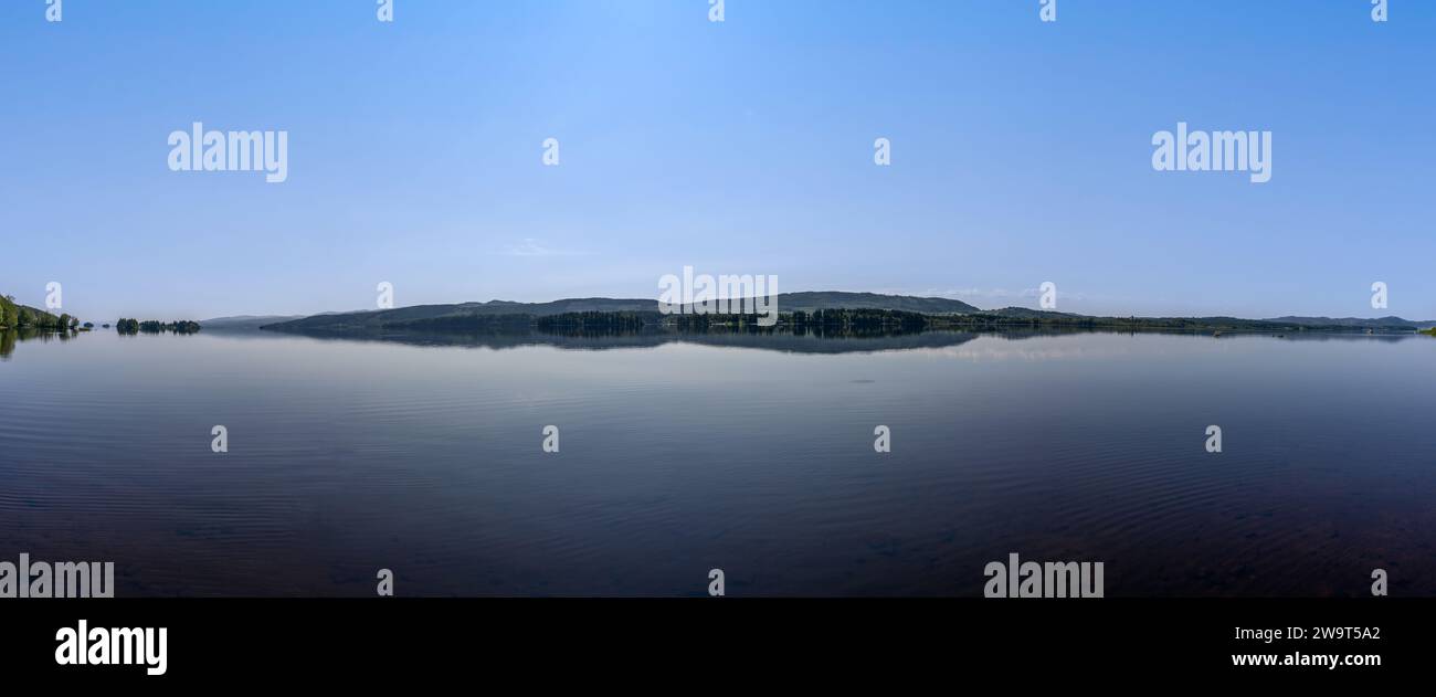 Panoramablick auf die Ufer des Snåsavatnet-Sees in Steinkjer, Norwegen, mit Bäumen, die unter einem leuchtend blauen Sommerhimmel auf dem Wasser reflektieren Stockfoto