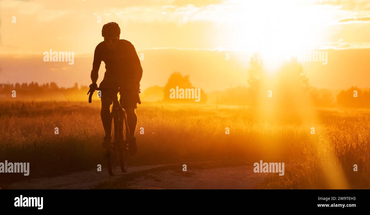 Silhouette eines Radfahrers auf einem Schotterrad, der auf einem Feld vor einem dramatischen Sonnenuntergang unterwegs ist. Stockfoto