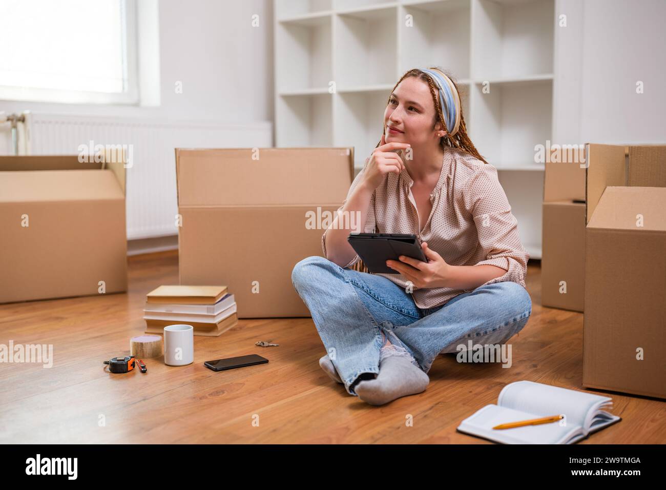 Moderne Ingwerfrau mit Zöpfen auf digitalem Tablet beim Einzug in eine neue Wohnung. Stockfoto