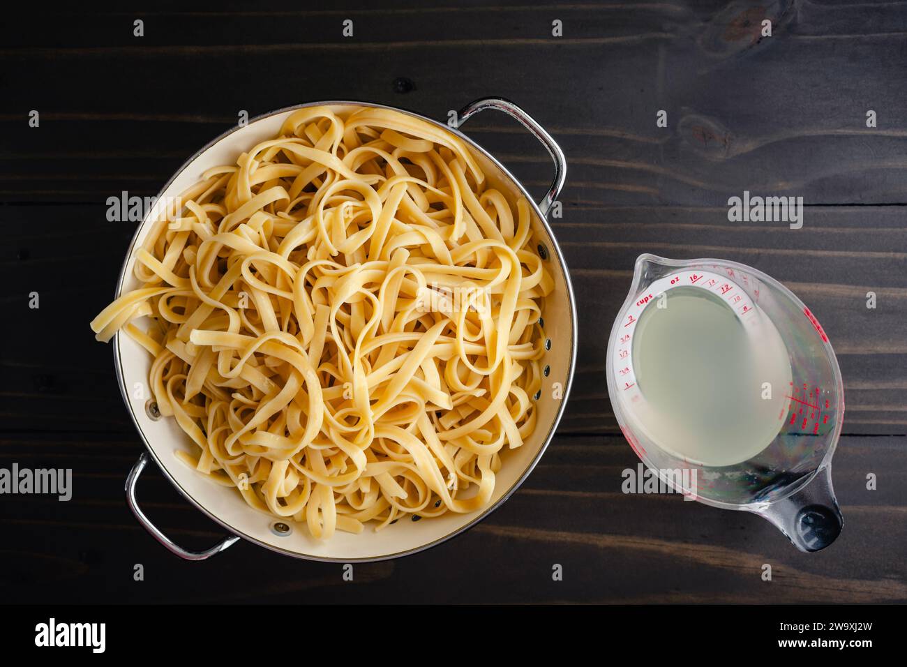 Gekochte Fettuccine in einem Sieb mit einer Tasse Nudelwasser: Gekochte und abtropfte Nudeln in einem Sieb mit einem Messbecher mit reserviertem Kochwasser Stockfoto