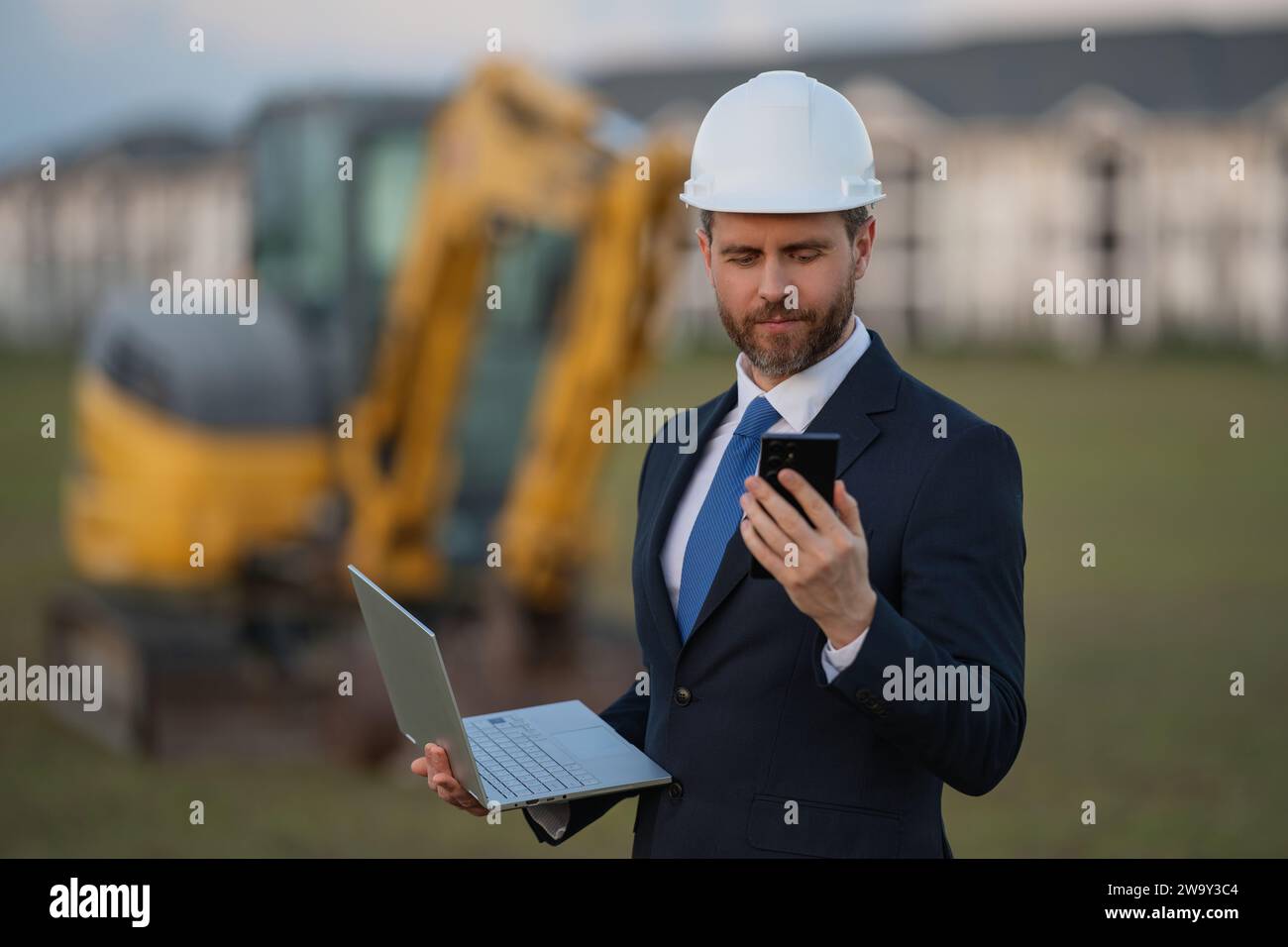 Baueigentümer in der Nähe des Baggers. Selbstbewusster Baubesitzer vor dem Haus. Architekt, Bauingenieur. Bauunternehmer mit Sicherheitsvorrichtungen Stockfoto