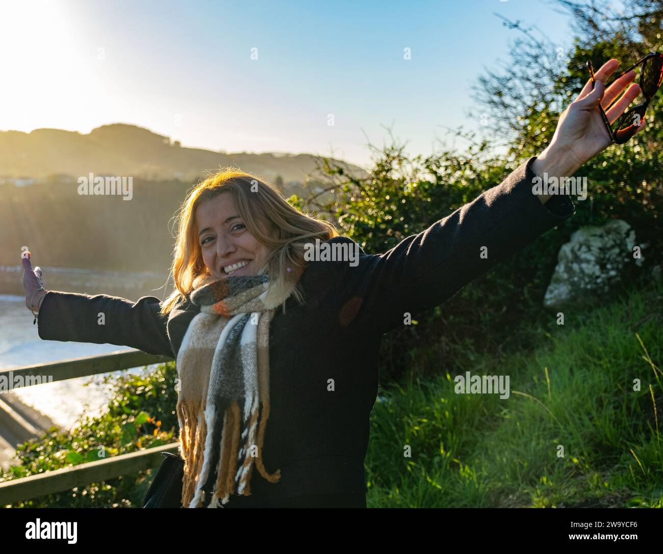 Porträt eines lächelnden blonden Mädchens, das eine Wolljacke und einen braunen karierten Schal trägt. Im Hintergrund des Bildes sehen Sie eine Küstenlandschaft wi Stockfoto