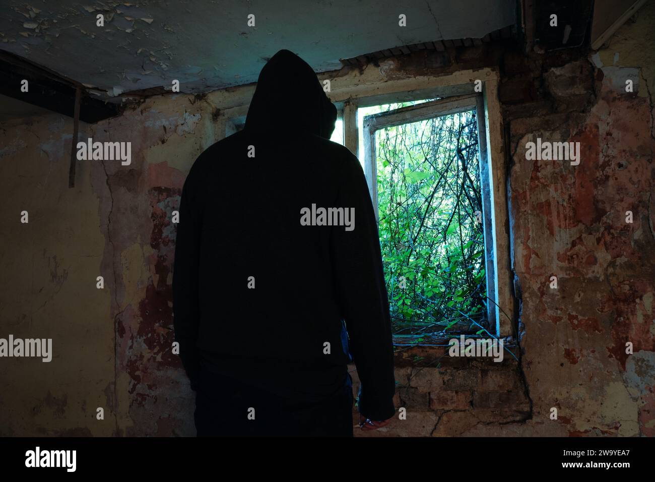 Ein Horror-Konzept einer gruseligen Kapuzenfigur, zurück zur Kamera. Silhouette durch ein Fenster in einem verlassenen Haus. Mit verfallenen Wänden. Stockfoto
