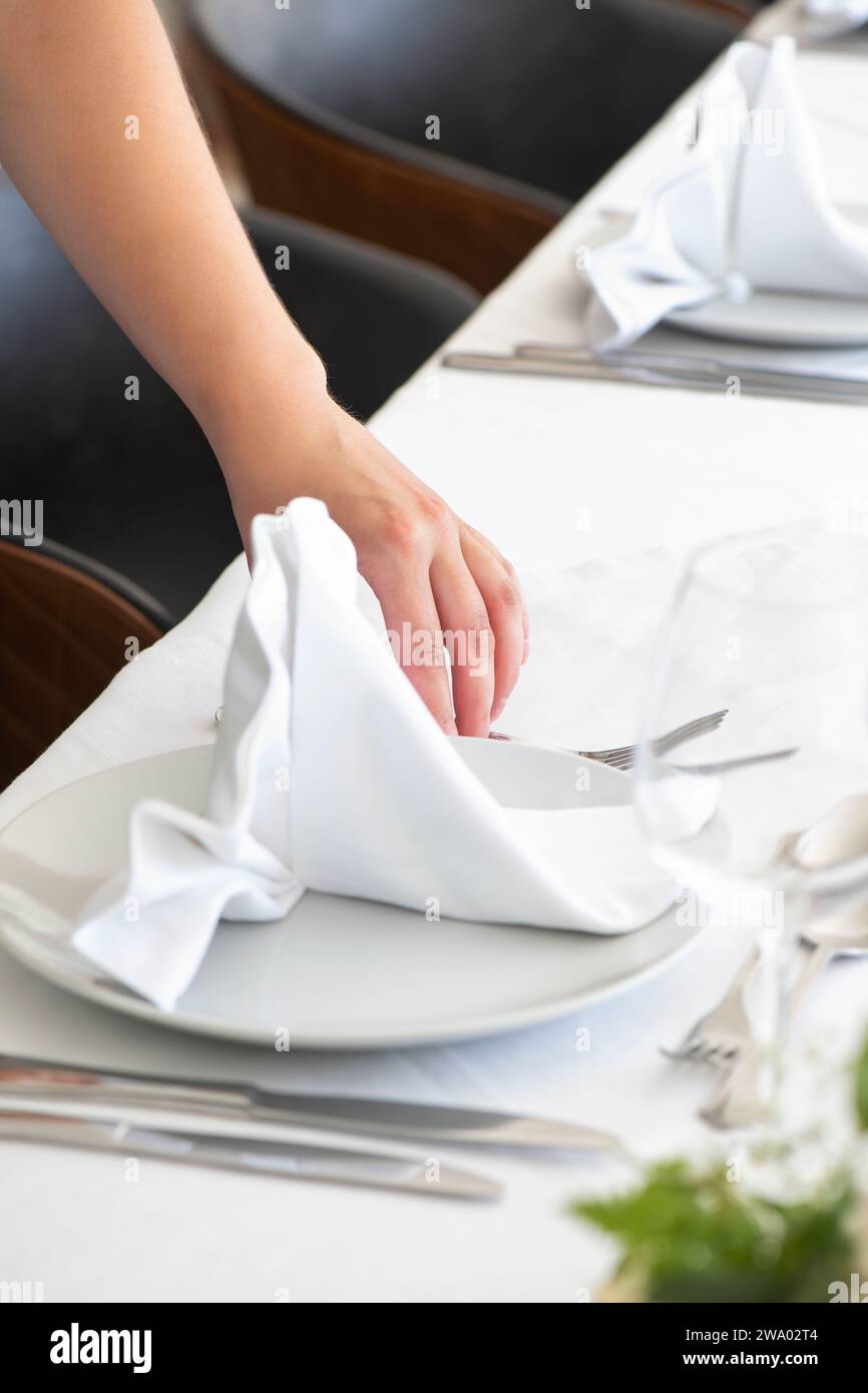 Die Damenhand faltet eine weiße Baumwollserviette zart, die dekorative Technik verleiht der Tischvorbereitung einen eleganten Touch und bereitet das Abendessen auf Stockfoto