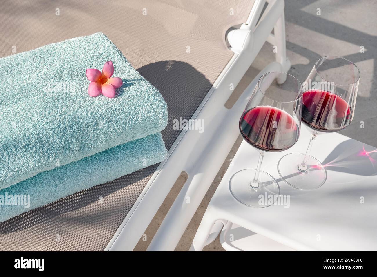 Sonnenliege mit zwei ordentlich gefalteten Handtüchern und einem weißen Tisch in der Nähe mit zwei hohen Gläsern Rotwein, einladende Lage am Pool, die eine Sommerstimmung verleiht Stockfoto