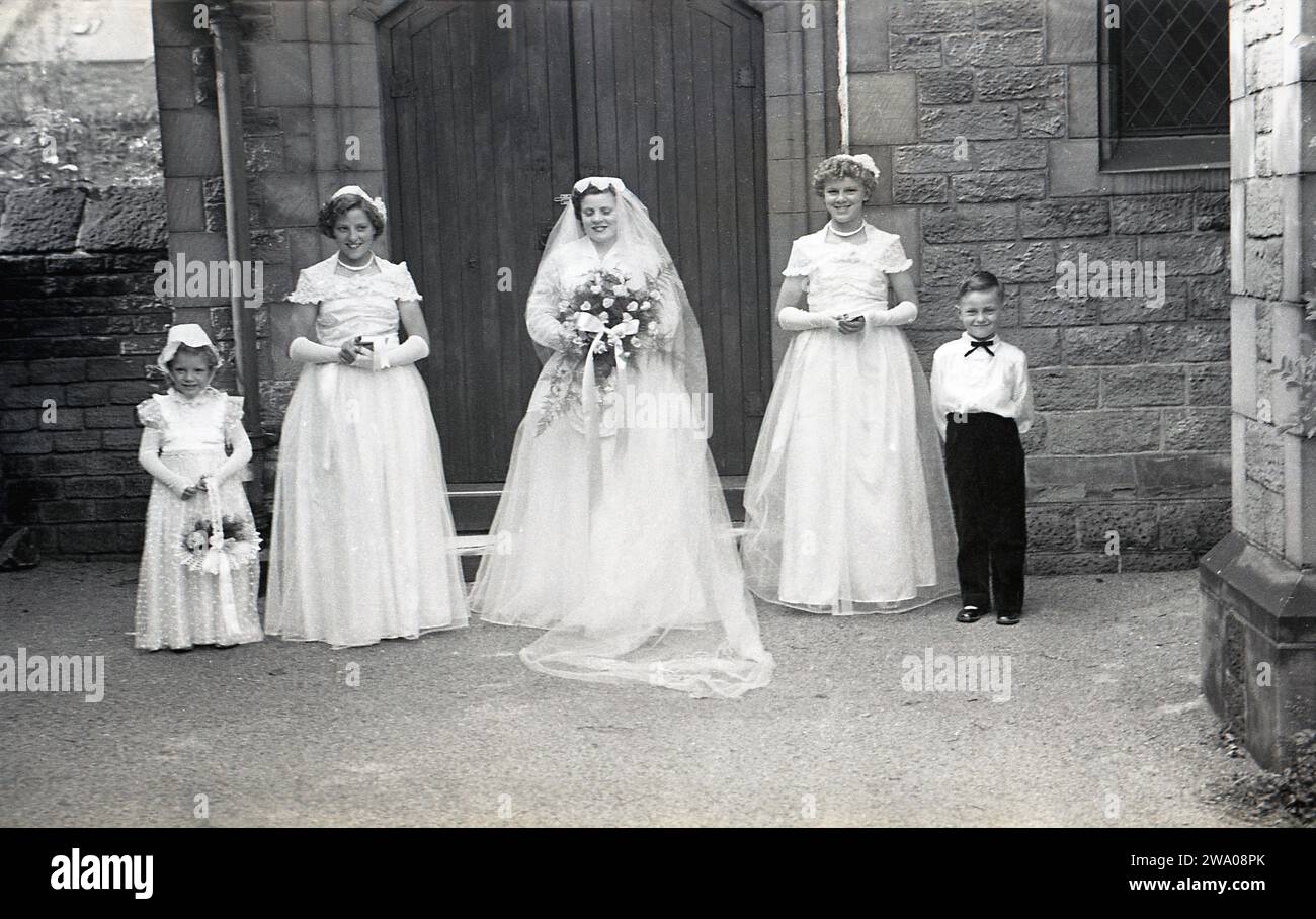1950er Jahre, historisch, Hochzeit, ein kleines Pageboy und ein junges Blumenmädchen, das für ein Foto mit zwei Brautjungfern und einer neuen Braut steht, außerhalb der Holztüren der Kirche, England, Großbritannien. Stockfoto