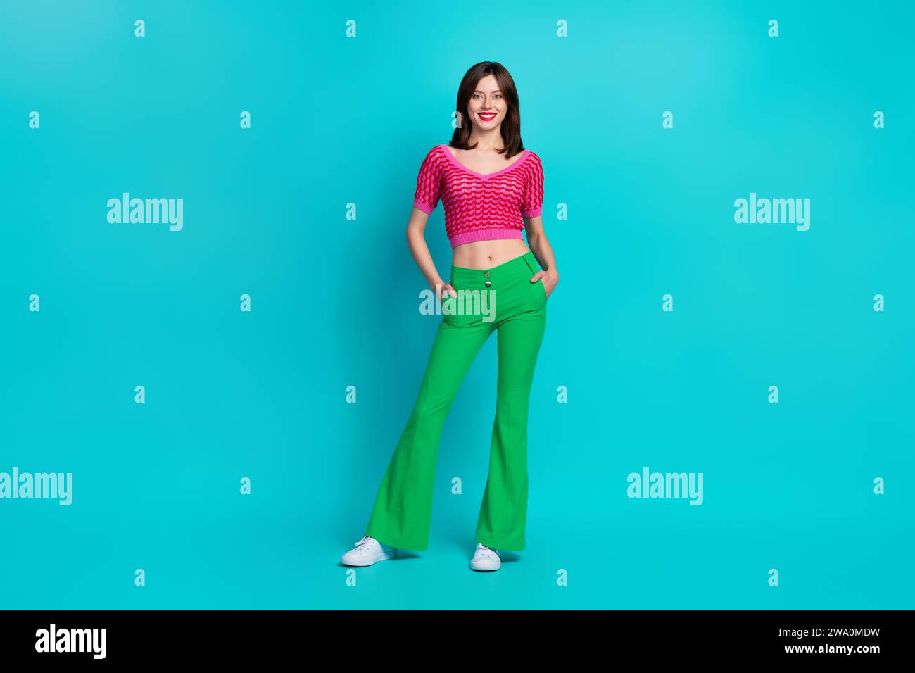 Foto in voller Länge von positiver fröhlicher Frau gekleidet Strickmode Oberteil grüne Hose hält Arme in Taschen isoliert auf blauem Hintergrund Stockfoto