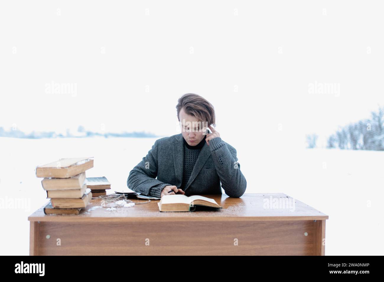 Ein junger, gutaussehender, intelligenter Mann in einer Jacke sitzt an einem Tisch auf einem Feld und liest im Winter alte Bücher Stockfoto