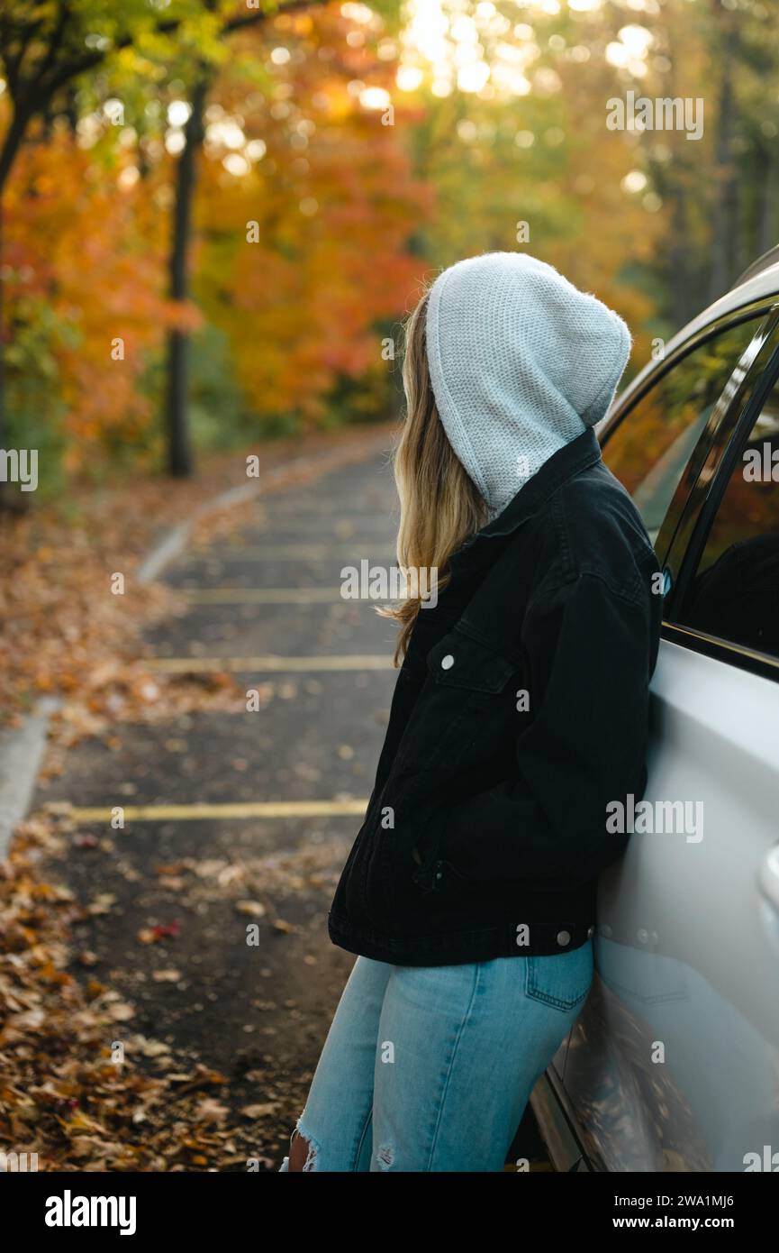Junge Frau, die sich auf ein Auto lehnt und auf dem Parkplatz Herbstfarben ansieht Stockfoto