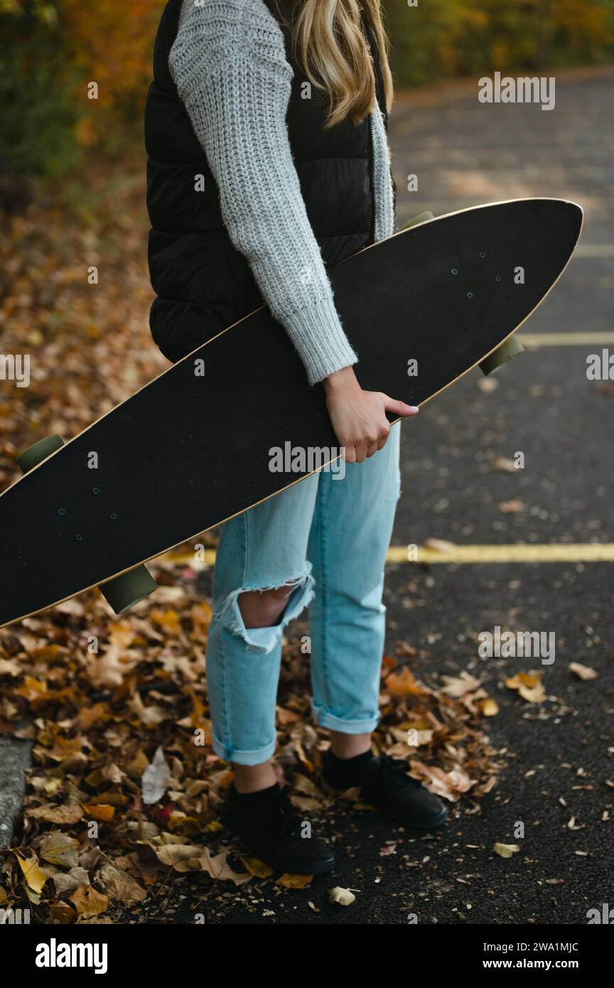 Zugeschnittene Ansicht einer Frau, die Longboard auf einem Parkplatz mit Blättern hält Stockfoto