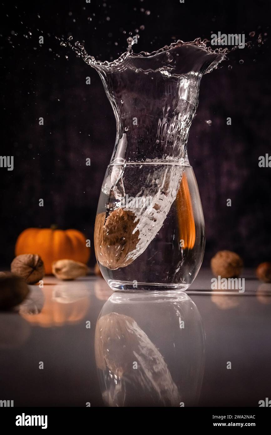 Spritzer Nussbaum in einem mit Wasser gefüllten Glas auf dunklem Hintergrund. Herbstzutaten im Hintergrund: Kürbis, Walnüsse, Mandeln usw. . Stockfoto