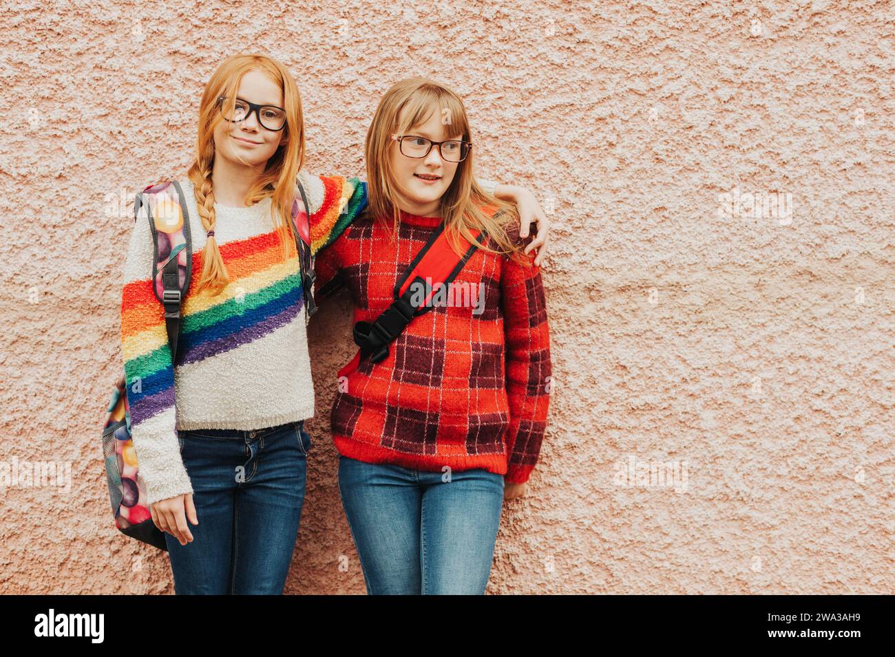 Gruppe von zwei entzückenden Mädchen, die draußen vor rosa Wänden posieren, Brille, Schulrucksäcke und bunte Pullover tragen, Back-to-School-Betrüger Stockfoto