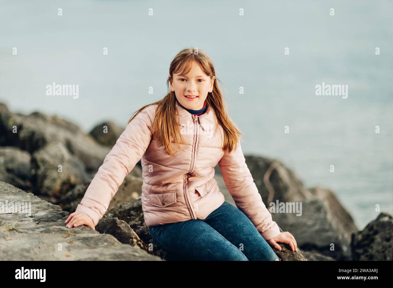 Niedliches, kleines 9-10-jähriges Mädchen, das an einem kalten Tag am See spielt und eine warme, pinkfarbene, gepolsterte Jacke trägt Stockfoto
