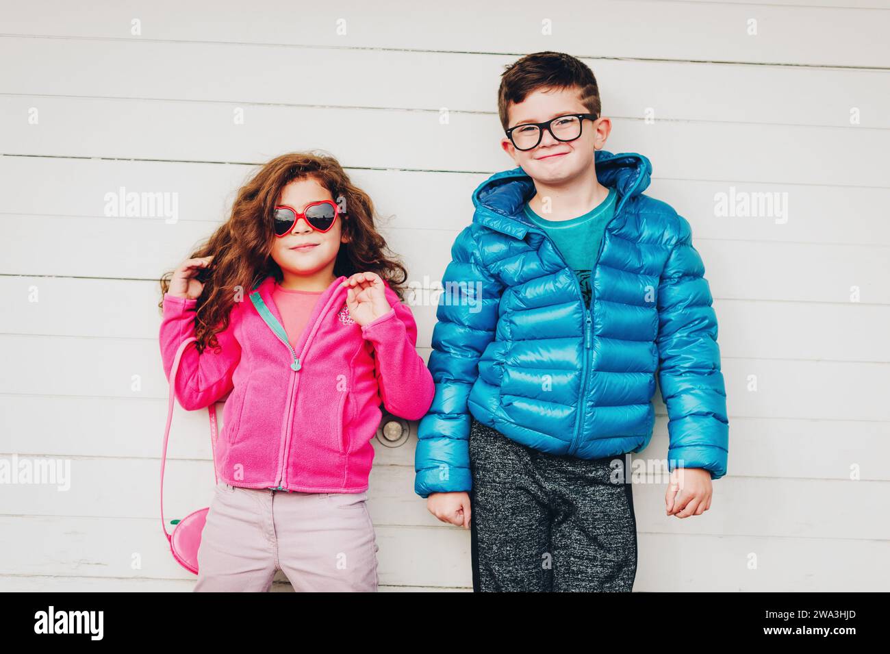 Das niedliche Kleinkind Mädchen und ihr großer Bruder posieren draußen vor weißem Holzhintergrund, tragen eine rosafarbene Fleecejacke und einen blauen wattierten Mantel Stockfoto