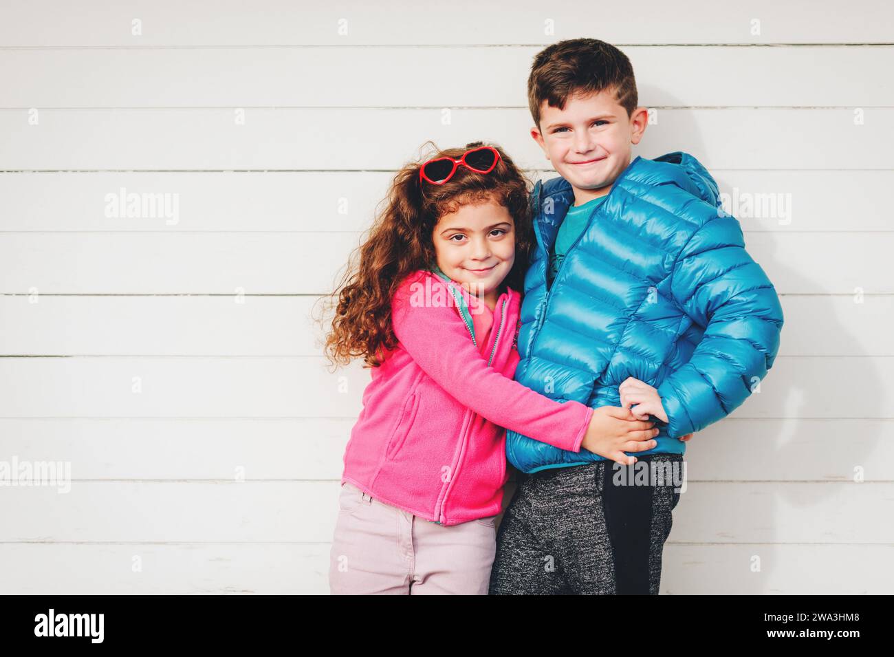 Das niedliche Kleinkind Mädchen und ihr großer Bruder posieren draußen vor weißem Holzhintergrund, tragen eine rosafarbene Fleecejacke und einen blauen wattierten Mantel Stockfoto