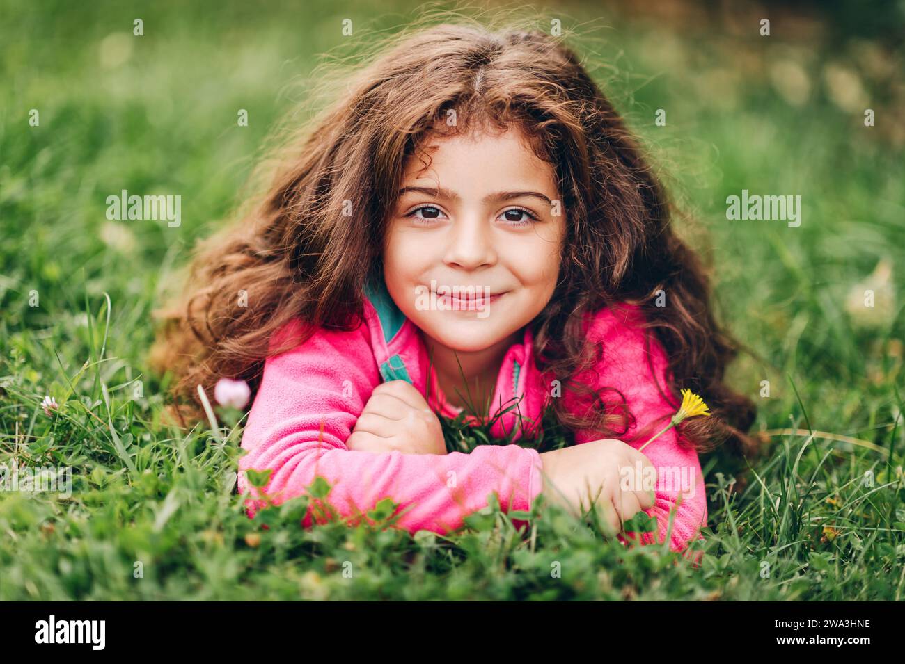 Outdoor-Porträt eines entzückenden kleinen 3-4-jährigen Mädchens mit schönen langen Haaren, in rosa Fleecejacke, auf grünem Gras liegend, mit gelbem Floß Stockfoto