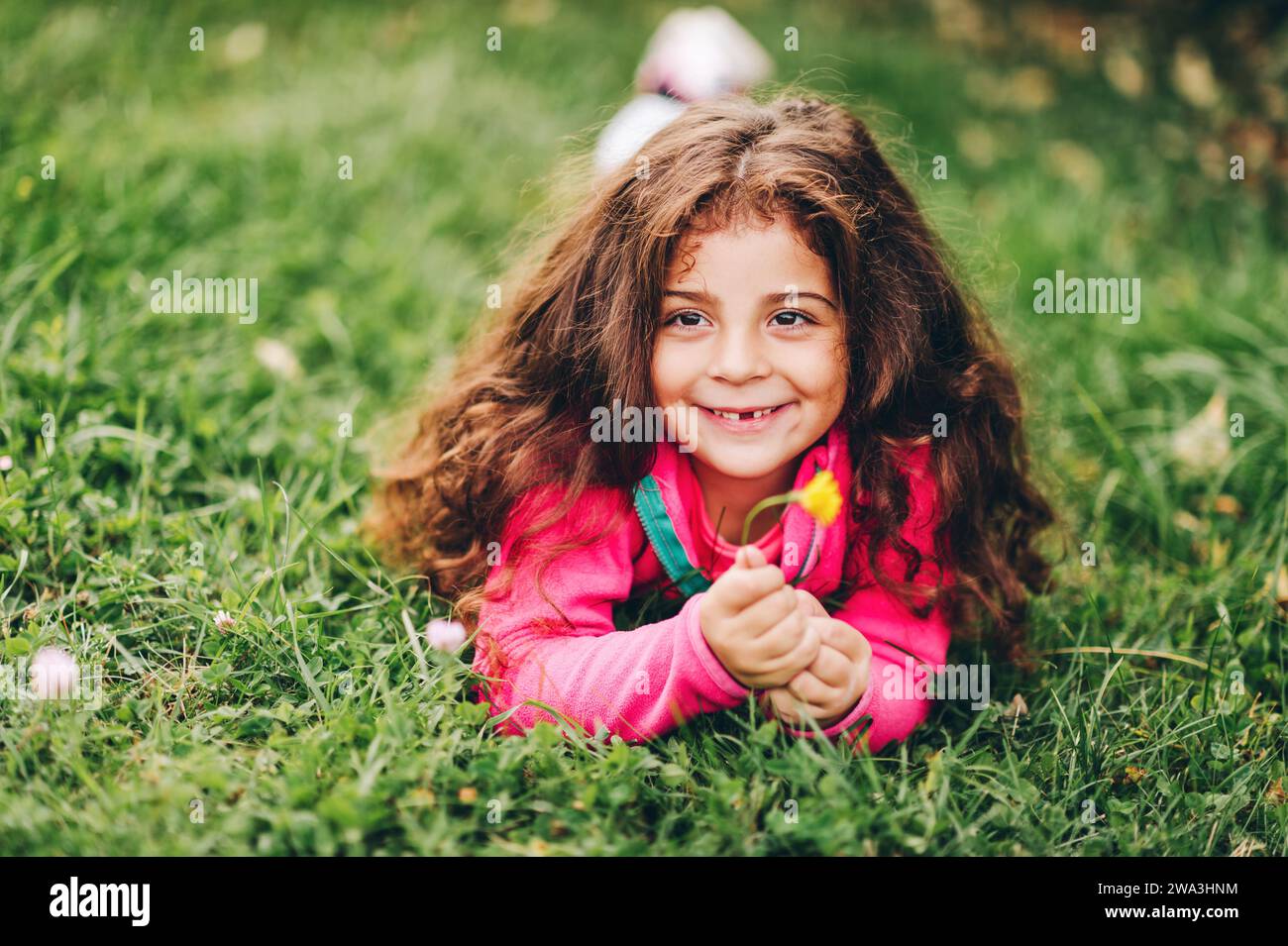 Outdoor-Porträt eines entzückenden kleinen 3-4-jährigen Mädchens mit schönen langen Haaren, in rosa Fleecejacke, auf grünem Gras liegend, mit gelbem Floß Stockfoto