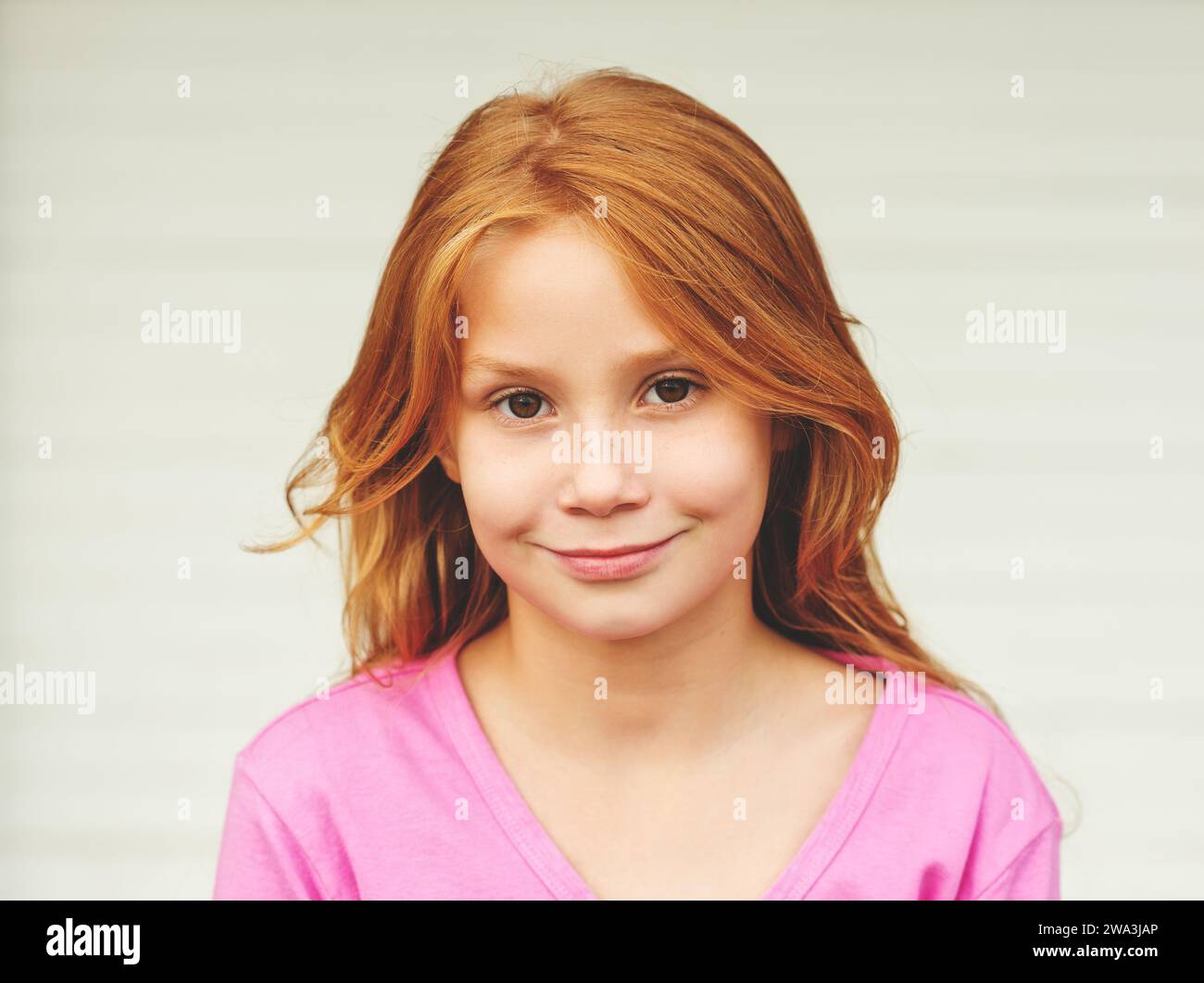 Außenporträt eines süßen kleinen 8-9-jährigen Mädchens mit langen roten Haaren, in rosa Jacke, stehend vor weißem Hintergrund Stockfoto