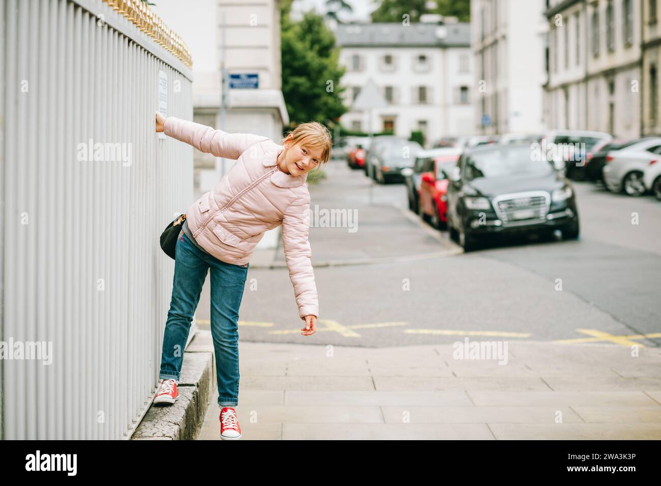 Außenporträt des lustigen kleinen Mädchens auf den Straßen von Genf, Schweiz. Das Kind trägt eine weiche pinkfarbene, gepolsterte Jacke, Jeans und rote Tennisschuhe Stockfoto