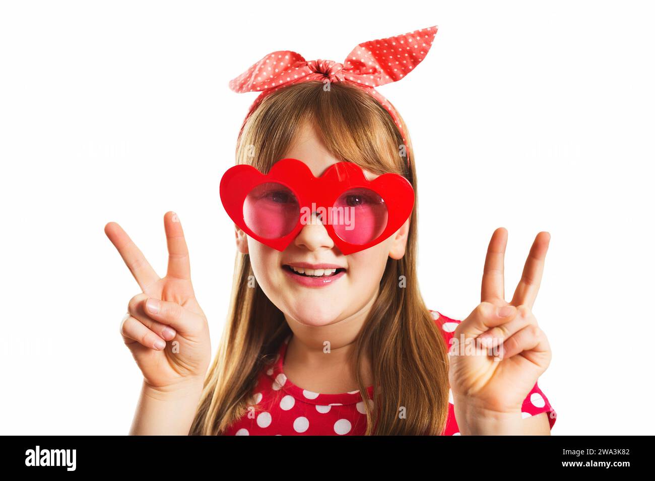 Lustiges Porträt eines süßen kleinen Mädchens mit großer herzförmiger Sonnenbrille und Polkadot-Stirnband, mit Finger Friedenszeichen, Studio-Aufnahme, isoliert auf W Stockfoto