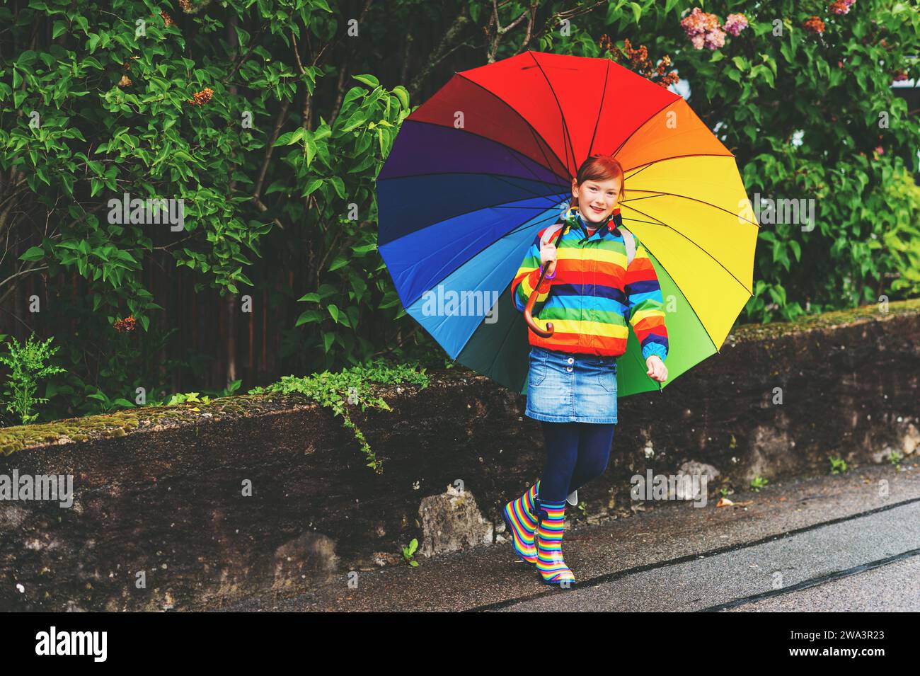 Lustiges kleines Mädchen, das draußen unter dem Regen spielt und einen großen bunten Regenschirm hält Stockfoto