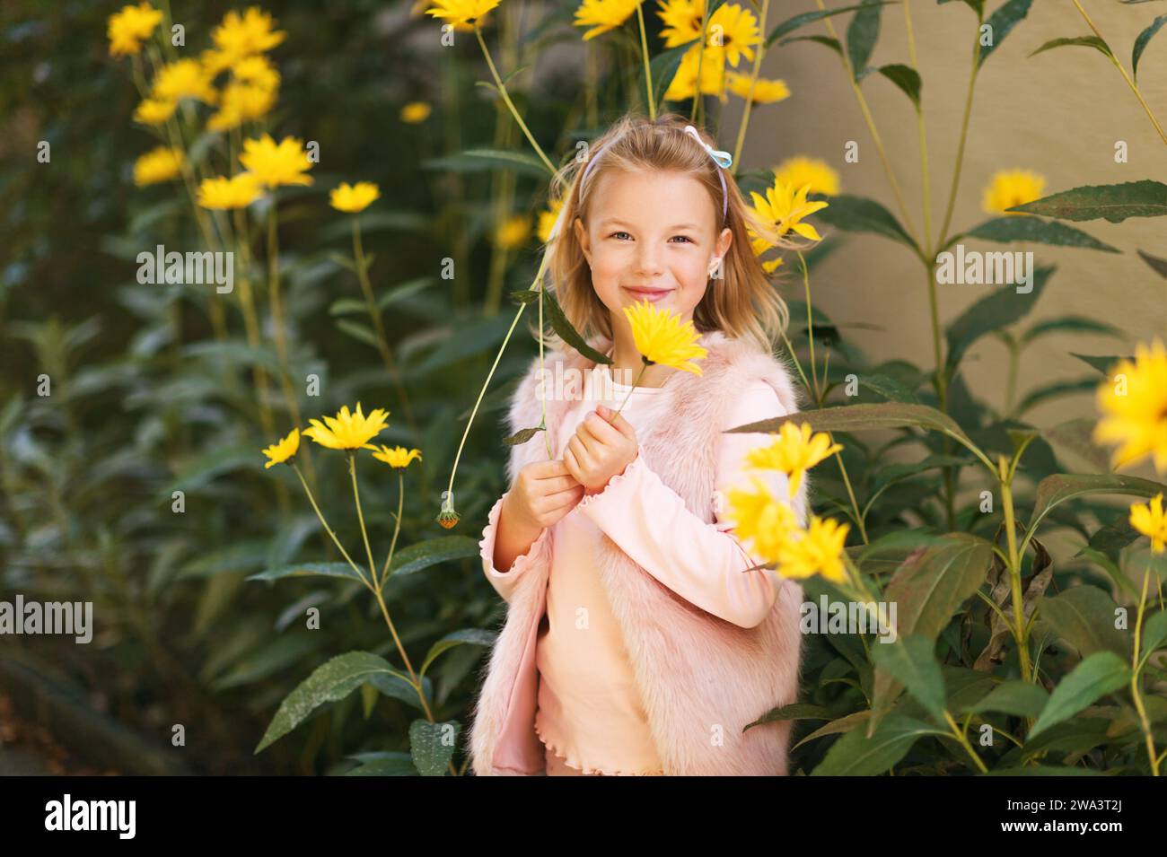 Außenporträt eines entzückenden kleinen Mädchens, das mit gelben Chrysanthemen-Blumen im Herbstgarten spielt, Kind mit hellrosa Kunstpelzjacke Stockfoto