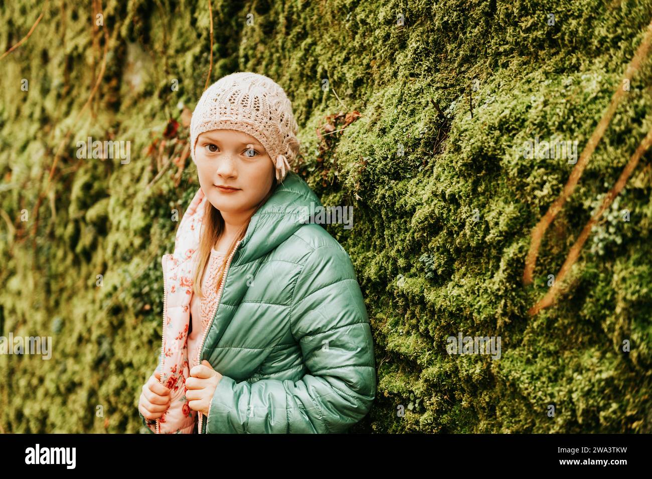Außenporträt eines süßen kleinen Mädchens mit rosa Hut und grüner Jacke, das neben der Mooswand posiert Stockfoto
