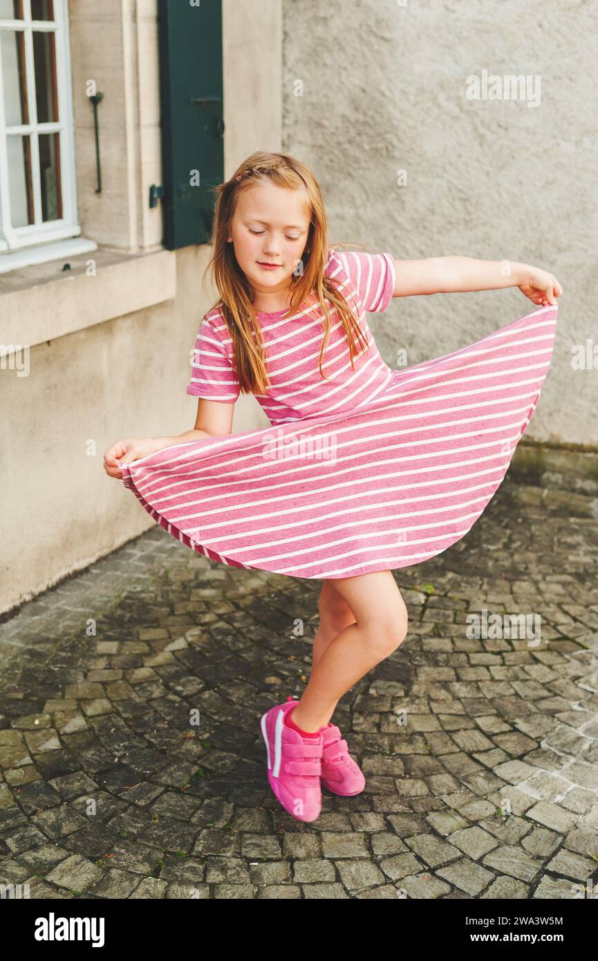 Outdoor-Porträt eines bezaubernden 6-jährigen Mädchens mit rosa Streifenkleid und Turnschuhen Stockfoto