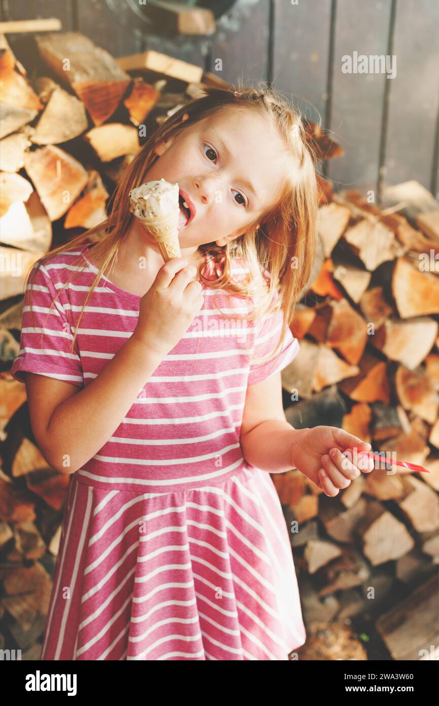 Süßes 6-jähriges Mädchen, das draußen Eis isst, rosa Streifenkleid trägt, Sommervergnügen für Kinder Stockfoto