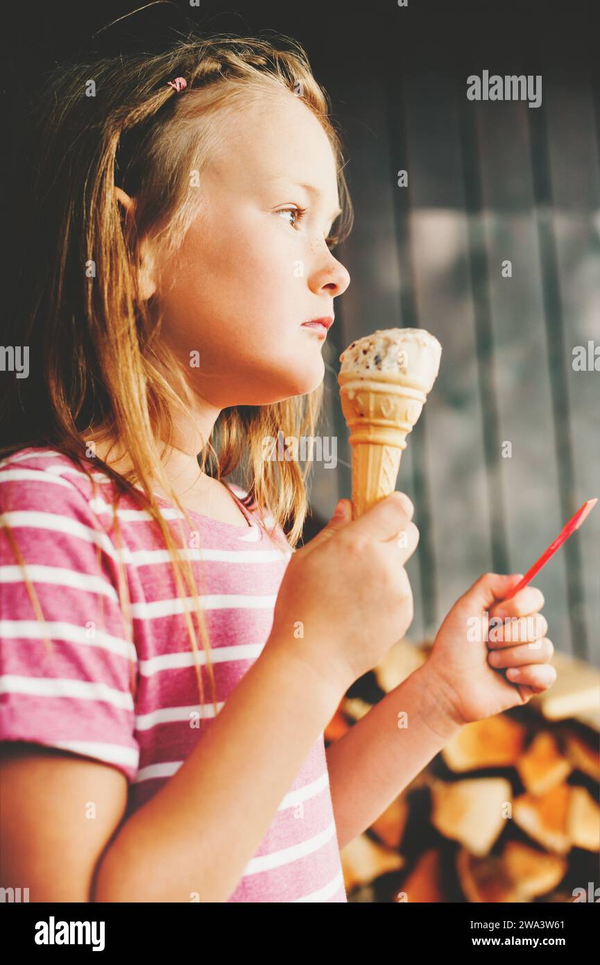 Süßes 6-jähriges Mädchen, das draußen Eis isst, rosa Streifenkleid trägt, Sommervergnügen für Kinder Stockfoto